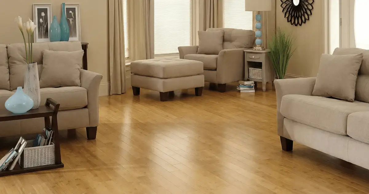 A living room with professionally installed bamboo flooring, beige sofas, a matching ottoman, a side table with a blue lamp, vases, framed pictures, and large windows with blinds and curtains in Bellevue, WA